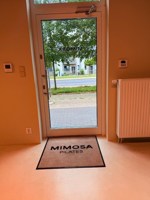 Beżowa mata z czarnym logo Mimosa Pilates przy przeszklonych drzwiach — realizacja BHP-Bejma